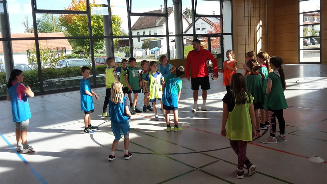 Kinder mit verschiedenfarbigen Leibchen stehen in der Mitte einer Turnhalle und ein Lehrer erklärt die Spielregeln.
