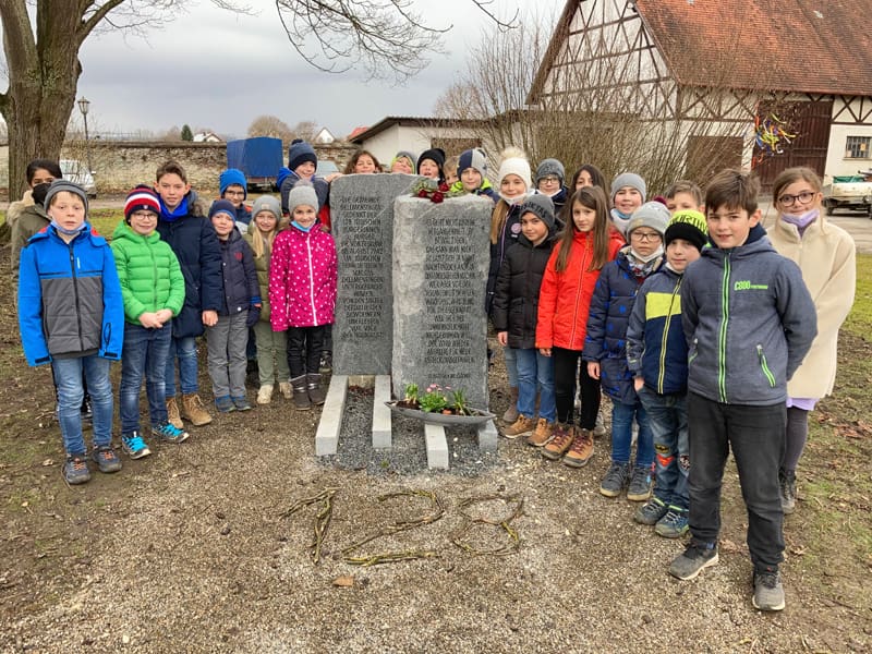 25 Schulkinder stehen um den Gedenkstein beim Schloss Dellmensingen. Vor dem Gedenkstein liegt die Zahl 128 aus Zweigen.