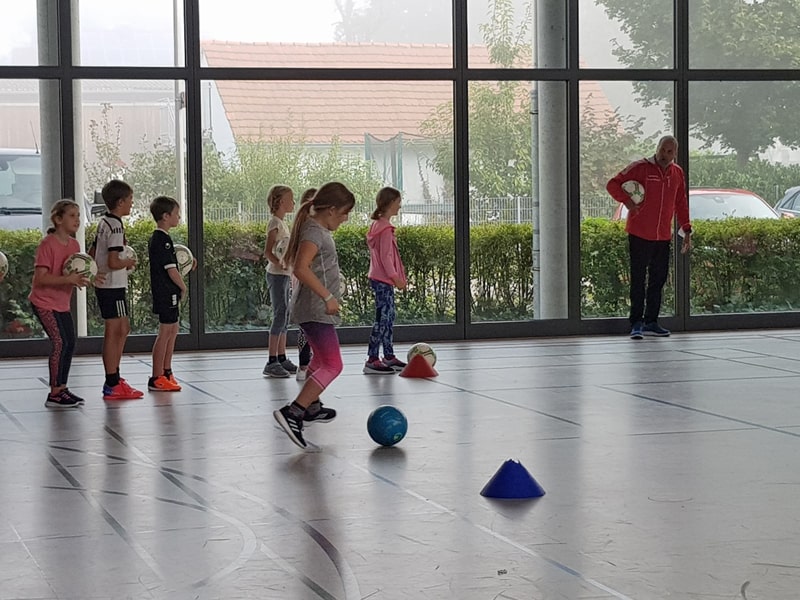 Kinder spielen in einer Turnhalle Fußball und ein Lehrer gibt Anweisungen.