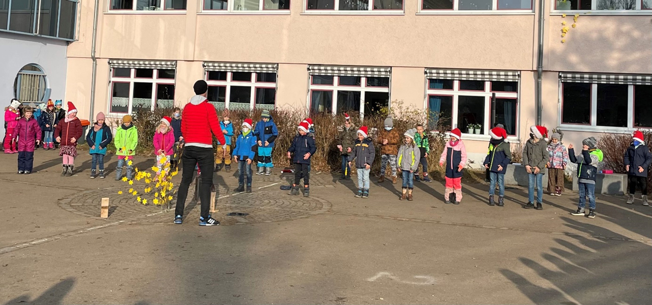 Warm angezogene Kinder zum Teil mit Nikolausmützen stehen auf dem Schulhof. Vor ihnen sieht man einen Lehrer von hinten.
