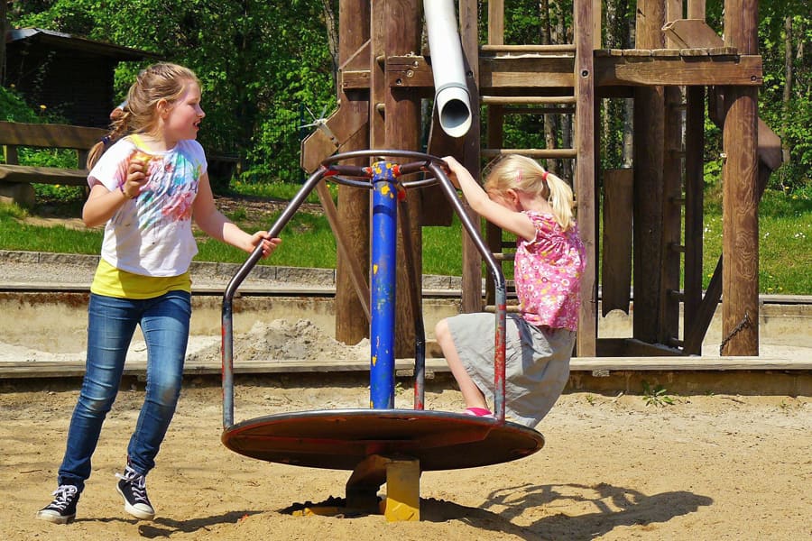 Zwei Mädchen spielen auf einem Spielplatz, ein Mädchen hockt auf einem Karusell und das zweite rennt um dieses.